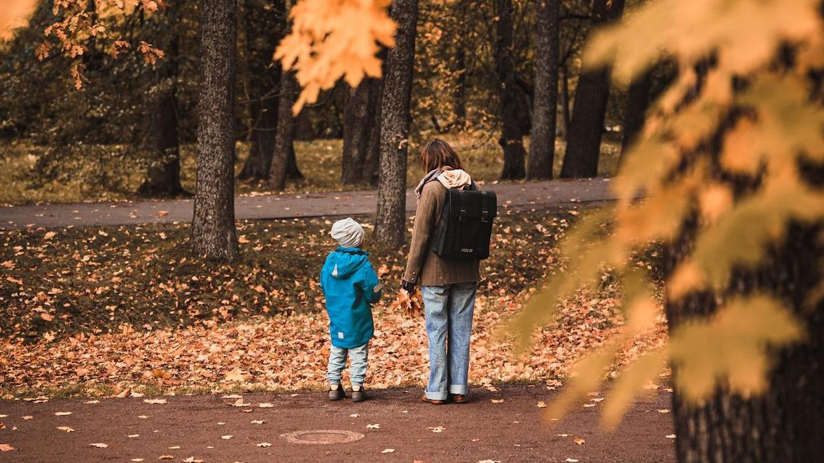 Un adulte et un enfant en manteau bleu, vus de dos, s’arrêtent au bord d’une allée de parc couverte de feuilles d’automne.